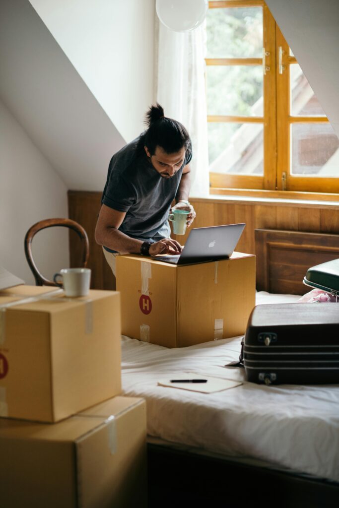A man with a ponytail using a laptop on a cardboard box while preparing to move in a bedroom.